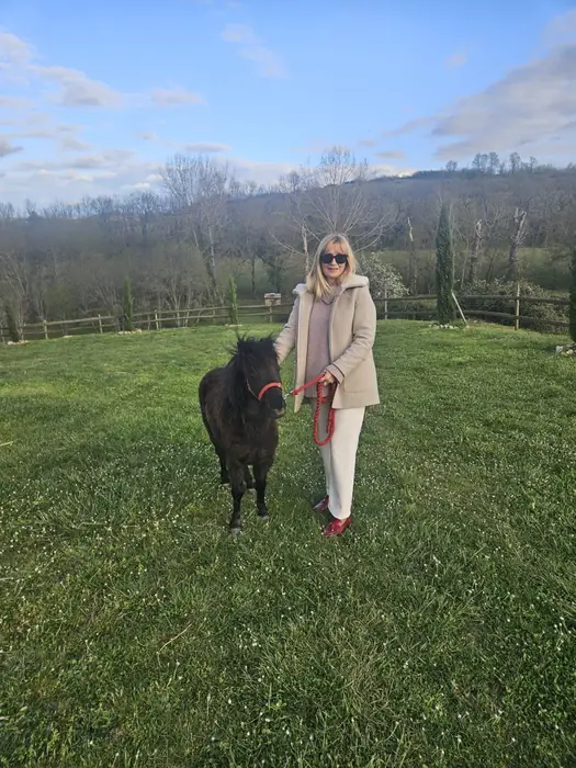 A woman in a cream coat and sunglasses standing on a sunny green lawn beside a small black Falabella on a lead.
